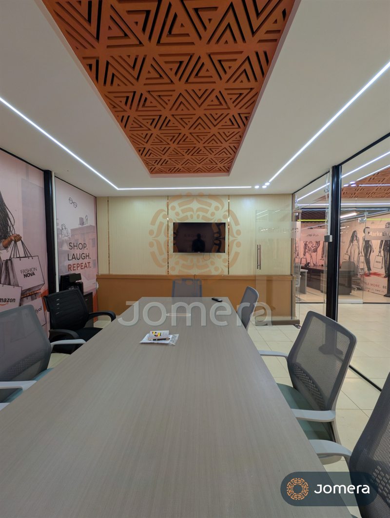 View down a boardroom table towards a TV wall, under a geometric CNC-cut ceiling panel and LED light lines
