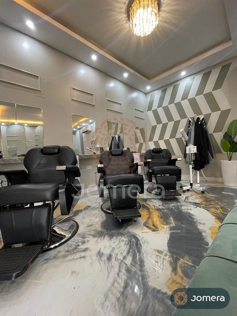 Row of black barber chairs facing brass-framed mirrors, with a crystal chandelier, wall mouldings and a sage chevron feature wall