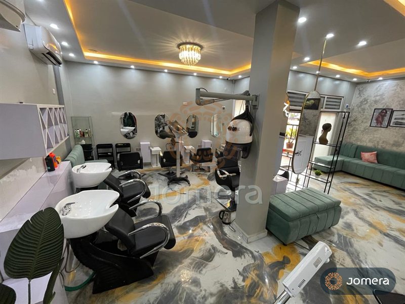 Salon wash basins and styling chairs with oval mirrors, a hood dryer, a sage velvet ottoman and lounge seating under cove lighting